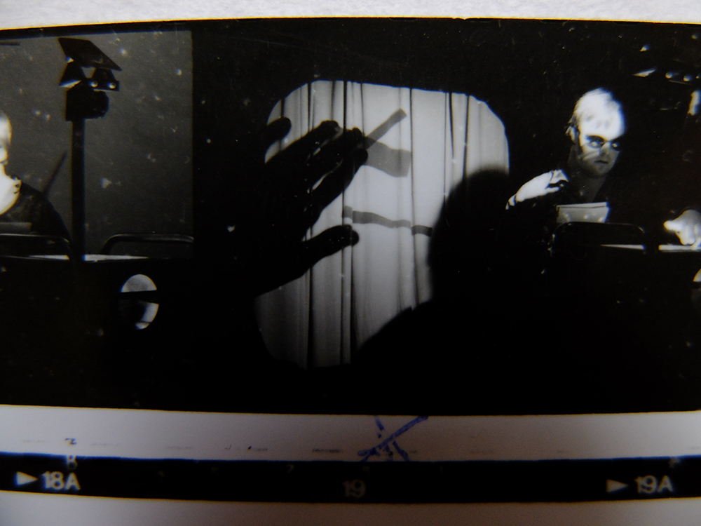 A black & white photo still of the performance Diary of a Dead Artist  by Corali Dance Company. The photo is taken in the original gallery of Southwark Park Galleries and features John Long  as an artist who creates an abstract picture projected behind him using an overhead projector.
